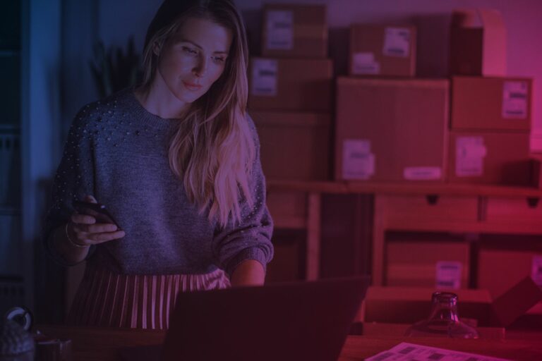 Frau steht mit Smartphone in der Hand vor Laptop, volle Kartons im Hintergrund